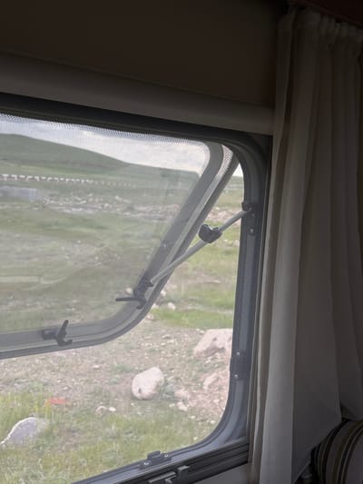 A close-up shot shows a window partially opened with a screen. The view through the screen reveals an outdoor scene in Tlik, Armenia, composed of a grassy field, sparsely dotted with rocks and patches of bare earth. The window is fitted with a mechanism to hold it open at various angles, with a white rod connected to the frame by black fixtures. Curtains are visible at the top of the frame, indicating the window is located inside a structure. The focus is on the window and its immediate surroundings, with the outdoor landscape providing context.