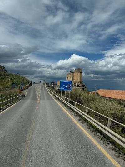The image depicts a two-lane asphalt road in Marina di Roseto Capo Spulico, Italy, under a dramatic, partly cloudy sky. The road, marked with faded white dashed center lines and solid yellow edge lines, recedes into the distance, appearing to ascend slightly and curve gently to the right. Light-colored metal guardrails line both sides of the roadway. On the right side of the road, a blue directional sign indicates routes to "TARANTO" straight ahead, "ROSETO C. SPULICO" to the right (towards the sea), and "castella" to the left. A smaller, circular speed limit sign displaying "40" is positioned near the blue sign.

Beyond the guardrail and the blue sign on the right, the multi-towered, light brown Castello di Roseto Capo Spulico is prominently situated on a rocky outcrop, overlooking the dark blue waters of the Mediterranean Sea visible in the far right background. Sparse palm trees are visible near the castle. Below the castle and closer to the foreground, a structure with a reddish-orange tiled roof is partially visible, surrounded by dense, tall, dry grasses. On the left side of the road, a grassy hillside slopes upwards, showing some patches of bare earth and indistinct small figures in the distance. The sky above features a mix of dark grey storm clouds and patches of bright blue. No distinct human activity or interaction is discernible in the foreground or midground.