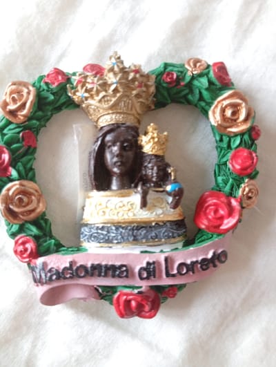 This media file contains a close-up, top-down shot of a religious figurine mounted on a heart-shaped wreath. The figurine depicts a dark-skinned Madonna holding a baby, both adorned with golden crowns. They are standing on a tiered base, with the Madonna wearing a yellow and white garment decorated with gold swirls and a dark grey, textured lower section. The heart-shaped wreath is made of green, ribbon-like material, possibly representing leaves and vines, and is adorned with several fabric roses in shades of red and gold. Some of the roses are realistic in appearance, while others are more stylized.
Below the figurine, a pink ribbon banner is attached to the wreath, displaying the text "Madonna di Loreto" in black, embossed lettering. The figurine and wreath are placed on a white, textured surface, likely fabric, which appears to be a tablecloth or a similar textile.
The lighting is bright, suggesting an indoor setting or outdoor daylight with diffused light, as there are no harsh shadows. There are no discernible environmental cues that indicate a specific time of day or weather conditions. The overall impression is that of a devotional item or souvenir. The presence of the text "Madonna di Loreto" suggests a connection to the Sanctuary of Loreto, a significant pilgrimage site in Italy. Given the location context of Civitanova Marche, Italy, this item could be a souvenir purchased from or related to the Loreto region.