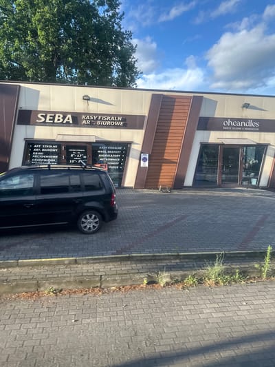 This is a medium-angle, exterior shot of a commercial building in Krosno Odrzańskie, Poland. The building has a modern design with a light-colored facade and brown architectural accents. Two businesses are visible: "SEBA" which sells cash registers and office supplies, and "ohcandles" which sells soy candles and home decor. A black minivan is parked in front of the SEBA store. The ground is paved with gray bricks. Tall green trees are in the background. The sky is blue with scattered white clouds.