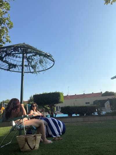 Two individuals are seated on lounge chairs on a grassy area next to a swimming pool. The person on the left, with long, dark hair, is wearing a bikini and facing away from the camera. The person on the right, with dark hair and sunglasses, is wearing a patterned bikini top and looking towards the camera. A large straw bag is on the grass in front of the person on the left. A blue and white striped towel is draped over the lounge chair of the person on the right. In the background, there is a swimming pool with people in it, green hedges, and buildings with red tile roofs. A large umbrella-like structure with foliage is present to the left of the frame, partially shading the scene. The sky is clear and blue. The location is identified as Villa del Prado, Spain.
