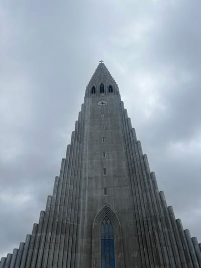 The Hallgrímskirkja church in Reykjavík, Iceland, is a tall, grey, concrete structure with a pointed spire. The church has a large clock on the front face. It is located at latitude 64.142 and longitude -21.927. The sky is overcast and the clouds are grey. There are no people visible in the image.