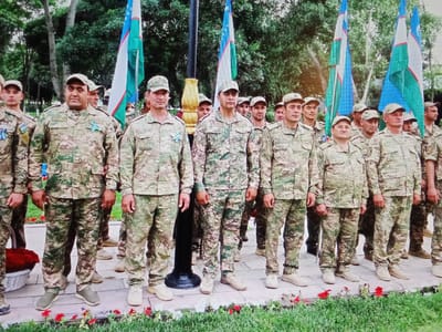 A group of approximately ten men in camouflage uniforms and caps stand in front of several flags. The flags appear to be national flags, with white, blue, and green colors, and a star and crescent motif. Some of the men have badges or ribbons on their uniforms, including a light blue ribbon on the uniform of the man in the center foreground. The men are standing on a paved walkway lined with grass and red flowers. Behind them, there are trees and foliage. The context suggests a formal gathering or ceremony, possibly military in nature, taking place outdoors. The image is taken from a slightly elevated perspective, looking down at the group. The overall lighting suggests daylight, possibly overcast. The man in the center foreground is facing the camera directly, while others are looking forward or slightly to the side. Some individuals are partially obscured by others or by the flags.