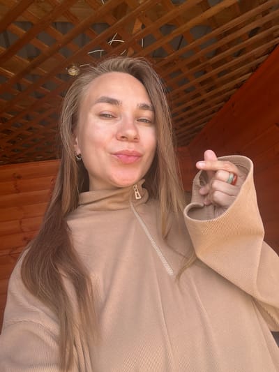 A young woman with blonde hair is taking a selfie outdoors. She is wearing a tan hoodie and has two rings on her right hand. The background consists of wooden beams and paneling, suggesting an outdoor structure like a gazebo or porch. There is no indication of the city of Arkadak, Russia in the image.