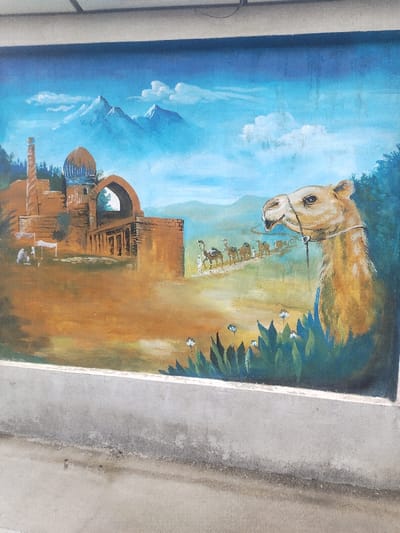 A vibrantly colored mural, painted on an exterior wall in Chighirik, Uzbekistan, depicts a panoramic Central Asian landscape. The mural features a large, light brown camel head in profile, facing left, prominent in the foreground on the right, with a rope bridle around its snout and green foliage below it. To the left, a complex of traditional brown-hued buildings stands, including a blue-domed structure with white script, a tall minaret-like tower, and a prominent arched gateway. Below these structures, two small figures and a white tent are visible. In the midground, a caravan of approximately seven to eight camels, led by two human figures, traverses a winding path from right to left. The background reveals a vast blue sky with white clouds and three snow-capped blue mountains. The ground of the mural transitions from brown to green, with small white flowers dotting the lower right. Below the painted surface, a concrete base, exhibiting minor discoloration and wear, forms the lower margin of the visible scene.