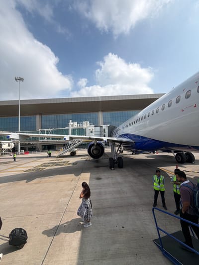 An aerial view shows an Indigo Airlines airplane parked at the terminal in Vikram Nagar, India, under a cloudy blue sky. The aircraft, with its distinctive blue and white livery, is connected to the terminal via a jet bridge. Several people are visible on the tarmac, including passengers and airline staff in high-visibility vests. One woman in a patterned dress stands on the tarmac, while a suitcase rests beside her. A staircase leads up to the airplane's entrance, and an orange traffic cone is placed nearby. The airport terminal building features a modern design with large glass windows. The scene is characterized by bright sunlight casting shadows across the concrete tarmac, marked with yellow lines indicating parking positions.