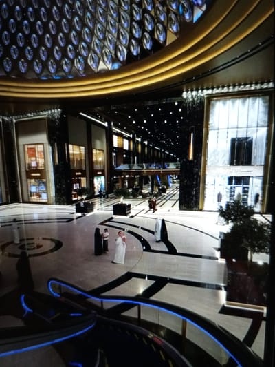 The image depicts an interior view of a large, modern shopping mall. The architecture features a grand, multi-level design with a prominent circular atrium. The ceiling is adorned with a decorative pattern of circular, textured elements, illuminated with blue light, and a golden, curved structure above. The mall's interior is spacious, with multiple levels visible, connected by escalators and walkways. The floor is a light-colored material with dark, geometric patterns.
Several individuals are present in the mall. In the foreground, a dark-colored escalator with blue lighting is visible. Further into the mall, on the ground level, three people are seen standing together, two in traditional Islamic attire (one in a black abaya and headscarf, the other in a white thawb and ghutra) and one person in lighter colored clothing. In the middle ground, several people are walking through the mall, some in groups. In the background, shops and store windows are visible, some with displays. The overall environment suggests a busy, upscale commercial space. There is no indication or possibility to determine the location in the city of Golbembal, Nigeria, as the image does not provide any identifying markers for that location.