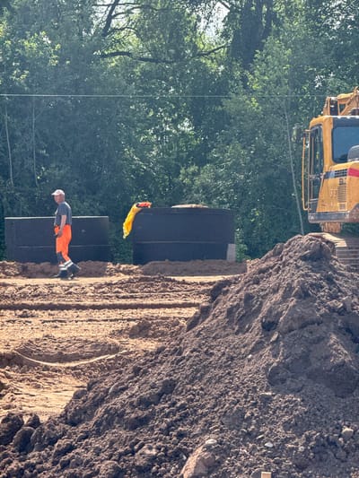 A man wearing an orange and blue work uniform walks across a dirt construction site in Kabanikha, Russia. He is wearing a light-colored baseball cap and black work boots. Behind him are two large, dark rectangular structures, possibly tanks or large pipes, stacked on top of each other. A bright yellow cloth is draped over the top of the right structure. To the right of the structures is a large yellow excavator. In the foreground, there are large piles of dirt, with some tire tracks visible in the sand. Lush green trees and foliage form the background of the scene, with a power line visible horizontally across the middle of the image.