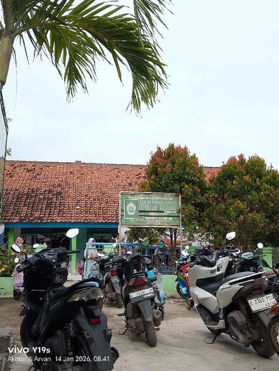 The image depicts a row of parked motorcycles in front of a building with a red tile roof. A sign identifies the building as "Taman Kanak-Kanak Uswatun Hasanah," which translates to "Uswatun Hasanah Kindergarten". Green leafy trees frame the scene. The sky is overcast. The photo was taken on January 14, 2026 at 08:41 with a Vivo Y19s phone by Akhtar & Arvan.