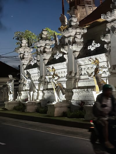 Captured at night in Kutuh, Indonesia, the photograph showcases a Balinese temple gateway. The architectural structure features intricate carvings of deities, statues, and traditional Balinese motifs. The gate is built from light-colored stone. Lush greenery and plants accent the architecture. A road with vehicular traffic is visible alongside the temple. A person is near the gate. The night sky serves as a backdrop. Electrical wires are near the structure.