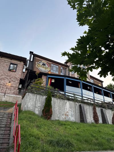 The image shows a view of a brick building with a large sign that reads "Bruges Brasserie". The building has multiple windows and is surrounded by greenery and concrete structures. A red railing is visible in the foreground, leading up a set of stairs. The scene appears to be outdoors, with a clear sky in the background. There are no people visible in the image. The time of day appears to be late afternoon or early evening, given the lighting. The weather seems to be mild and clear. The presence of the "Bruges Brasserie" sign suggests it is a restaurant or bar. The overall setting is a somewhat industrial or urban environment, with a mix of brickwork, concrete, and vegetation.
