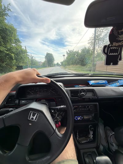 An interior view from the driver's seat of a car, likely a Honda Civic, is captured as the vehicle travels down a rural road in Kumachevo, Russia. The driver's left hand is on the steering wheel, which features the Honda emblem. The dashboard displays instruments, including a speedometer and tachometer, with red trim accents. A car stereo is visible below the air vents. On the passenger side of the dashboard, a package of Oreo cookies is placed. Above the windshield, a rearview mirror is mounted, with two black cartoon-like decorations hanging from it, one with "Top G" text. Outside the car, trees line both sides of the asphalt road, with houses and a wooden fence visible in the distance. The sky is partly cloudy with cirrus formations. The image composition suggests it was taken by a passenger or by the driver using a wide-angle lens or selfie mode.