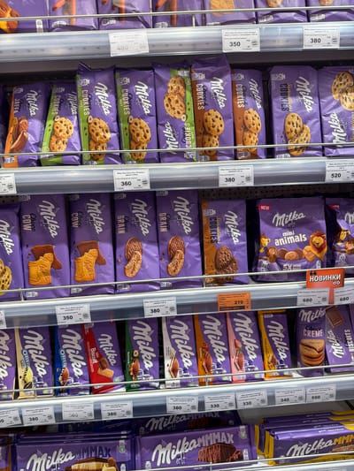 This media file is a close-up shot of a refrigerator display filled with various Milka brand chocolate and cookie products. The primary objects are rows and stacks of individually packaged Milka snacks, predominantly in the brand's signature purple packaging. Visible product types include cookies, biscuits, and chocolate bars with different flavor variations such as "Choco Nut," "Choco Cow," "Sensations Cookies," "Choco Grains," "Mini Animals," "Crème & Biscuit," "Strawberry," "Caramel," "Yoghurt," "Biscoff," and "Oreo." Some packages display images of the specific product inside, like cookies with chocolate chips or embossed chocolate bars.
There are no people or explicit scenes depicted, nor any indication of the time of day or weather. The setting is clearly a retail environment, likely a supermarket or a convenience store, indicated by the refrigerated display unit and the organized arrangement of products. The overall impression is one of abundant consumer goods presented for sale.
Visible text is extensive due to product labeling. Prominent text includes the "Milka" logo in its distinctive font. Many packages feature descriptions of the product, such as "Cookies," "Biscuits," "Choco," and flavor names like "Nut," "Strawberry," and "Caramel." Nutritional information, ingredient details, and brand taglines like "With Alpine Milk" are also present. Additionally, price tags and labels with product codes and weights are visible on many of the items, with some prices like "520," "380," "350," "360," "270," "680," "590," "340," and "330" being legible. Some labels also include the text "SWEET LAVKA" and Cyrillic script. There's also a prominent "НОВИНКИ" label, meaning "New Arrivals" in Russian.