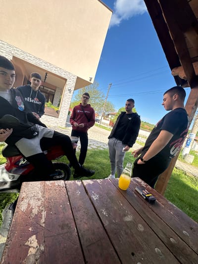 A group of young men is gathered around a wooden picnic table outside, likely in Lenovo, Bulgaria, based on the query. One man is leaning over the table, looking down. Another man is standing next to him. On the table are a pack of cigarettes and a glass bottle filled with yellow liquid. Two other men are standing or sitting further away from the table. One of them is sitting on a red and black scooter or motorcycle. A building is visible in the background. The sky is blue and sunny.