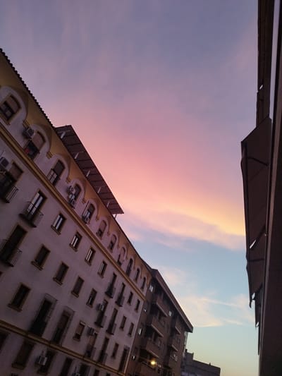 The image captures a city scene in Jerez, Spain, during twilight. The sky displays a gradient of soft pinks, purples, and pale blues, indicative of sunset or sunrise. Dominating the left side of the frame is a multi-story building with a light-colored facade, adorned with arched windows and balconies. Several air conditioning units are visible on the exterior of this building. To the right, another building is partially visible, appearing darker and more modern in its construction. The perspective is angled upwards, emphasizing the sky and the architectural elements of the buildings. No people are present in the frame, and the scene is still and serene.