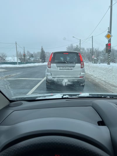 The image captures a snowy winter road scene in Voronino, Russia, from the perspective of a vehicle stopped behind another. In the midground, a silver SUV is stationary on an asphalt road partially covered in snow and slush. The rear of the SUV is soiled with road grime and snow, displaying a Russian license plate "E 353 OA 50". Its red tail lights are illuminated, and a snow-covered tow hitch is visible below the bumper. The vehicle is positioned at an intersection, indicated by a clear stop line on the road. To the right, a utility pole supports a red traffic light and a yellow diamond-shaped 'priority road' sign. Another red traffic light is visible further down the road on the left. The roadside features substantial snow banks, and the background consists of snow-covered bare trees, evergreen conifers, and distant structures under an overcast sky. Utility poles with overhead lines traverse the scene. The foreground shows a partial view of the observer's vehicle dashboard and steering wheel, with snow resting on the interior base of the windshield, which exhibits a visible crack. No individuals are present in the image.