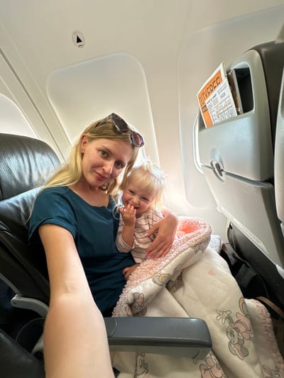 A blonde woman and a baby are seated on an airplane. The woman is wearing a dark teal shirt and sunglasses are perched on her head. She is holding the baby, who is wearing a striped shirt and is wrapped in a pink and white blanket with Mickey Mouse patterns. The baby is smiling and has its fingers near its mouth. The airplane cabin is visible, with seats and a safety card in a pocket on the seat in front. The image appears to be taken in Yehud Monosson, Israel.