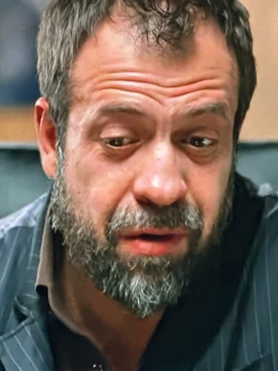 The image is a close-up, medium shot of a man's face. He has a salt-and-pepper beard and mustache. His hair is dark and appears short. His eyebrows are furrowed, and his eyes are looking downwards. He is wearing a dark pinstriped shirt or jacket over a lighter-colored collar. The background is out of focus and appears to be an indoor setting with muted colors. There is no visible indication of the city of Dondușeni, Moldova.