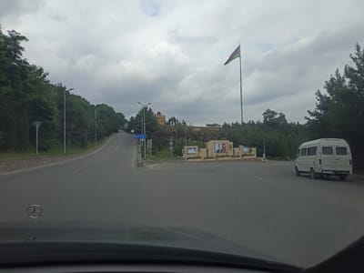 The image shows a road with a white van driving on the right side. In the background, there is a monument with portraits of two men and a flag of Azerbaijan. The monument is located in the city of Qusar, Azerbaijan, which is known for its mountainous terrain and beautiful natural scenery. The sky is cloudy, and the trees are lush green, suggesting it is summertime. A Mercedes-Benz logo is visible on the hood of the car from which the photo was taken.