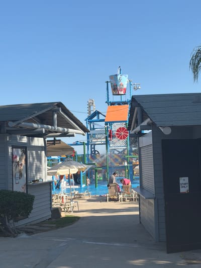 In Trachoni, Cyprus, this image captures a water park on a clear, sunny day. In the foreground, two grey, shed-like structures with peaked roofs partially obscure the view of a bustling water park. The structure on the left has a large poster depicting ice cream on its side, and a sign that reads "STAFF ONLY". The structure on the right is darker and also has a "STAFF ONLY" sign.
Beyond these structures, a vibrant water park unfolds. A large, colorful water play structure dominates the background, featuring slides and a tipping bucket that is actively showering water onto people below. Several individuals are visible interacting within the water park, some sitting on lounge chairs, others standing near the water. Beach umbrellas provide shade, and lounge chairs are scattered around the area. The ground is paved, and a clear blue sky serves as the backdrop, with a hint of a palm frond visible on the far right. The overall scene is one of a lively recreational environment.