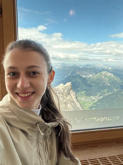 A young woman with brown hair and blue eyes smiles at the camera, her face filling the left side of the frame. She is wearing a light-colored jacket over a white turtleneck. Behind her, a large window offers a panoramic view of the Alps, with lush green valleys dotted with small villages and rugged, rocky peaks reaching towards a bright blue sky with scattered clouds. The text "Silvrettagruppe" is visible on the window, indicating the mountain range. The scene suggests the woman is at a high vantage point, likely a tourist attraction or a mountain hut in the Austrian Alps, possibly near Ehrwald.