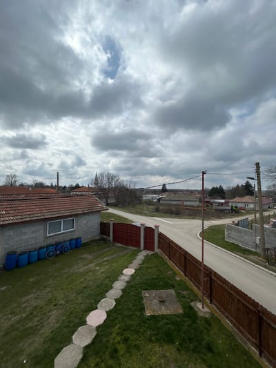 An elevated view depicts a residential street scene within the village of Sredkovets, Bulgaria, under an overcast sky with a singular patch of blue visible overhead. In the immediate foreground, a meticulously maintained grass lawn occupies the lower left quadrant. A linear path of approximately eight light-colored, octagonal stepping stones traverses the lawn from the bottom-center towards a closed, two-panel red metal gate positioned centrally. The gate is flanked by white-painted posts with reddish caps and opens onto an asphalt road. Adjacent to the right side of this property, a dark brown wooden fence extends horizontally, delineating the boundary from the roadway. Embedded within the lawn near the bottom-right is a square concrete slab featuring a circular metal cover in its center.

To the left of the lawn, a single-story structure constructed from grey concrete blocks features a reddish-brown tiled roof and a white-framed horizontal window. Lined horizontally against the base of this building are approximately nine blue plastic barrels. A single blue bicycle is parked leaning against the building, positioned between the barrels and the window.

The asphalt road, grey in color, forms a "Y" or "T" intersection, curving towards the right foreground and extending into the middle distance. A reddish-brown utility pole stands near the red gate, with power lines extending diagonally across the scene. A second utility pole with a street light is visible further down the road to the right.

Beyond the road and immediate properties, numerous residential buildings with varied rooflines and construction materials are scattered across the midground and background. Predominantly bare deciduous trees, indicative of late winter or early spring, are interspersed among the structures. A tall, white church spire is distinctly visible above the treeline in the left-center distance. To its left, a slender, dark industrial chimney or stack protrudes vertically. The horizon is hazy, blending into the consistent grey of the cloudy sky. No individuals or animals are present within the frame.