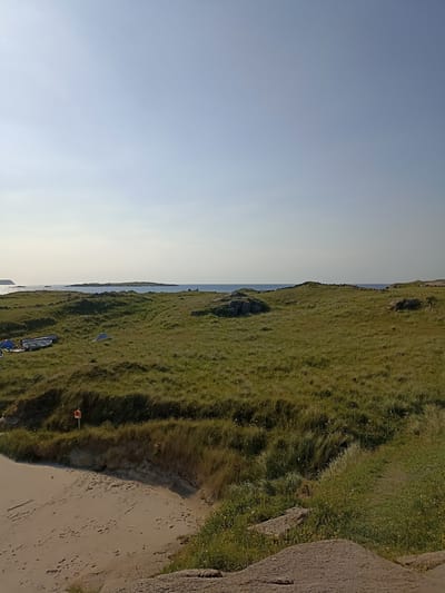 The image depicts a coastal landscape with grassy dunes leading to the sea. In the foreground, a sandy beach is visible, with evidence of erosion on the dune face. Several boats and at least two tents are situated on the grass to the left, suggesting a camping or recreational area. A red and white marker sign stands on the edge of the dune. In the distance, islands are visible in the calm, blue sea under a clear, light blue sky. The scene appears to be set in Carnboy, Ireland, characterized by its rugged coastline and natural beauty.