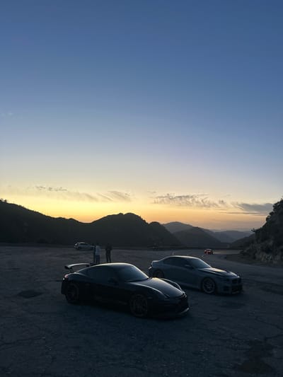 Two sleek, dark-colored sports cars sit parked on an asphalt surface at dusk. Mountains form the backdrop, silhouetted against a fading orange and yellow sky. A few people stand near the cars. In the distance, another car can be seen further up the road. The image shows a scenic overlook with the gathering darkness accentuating the contours of the landscape.