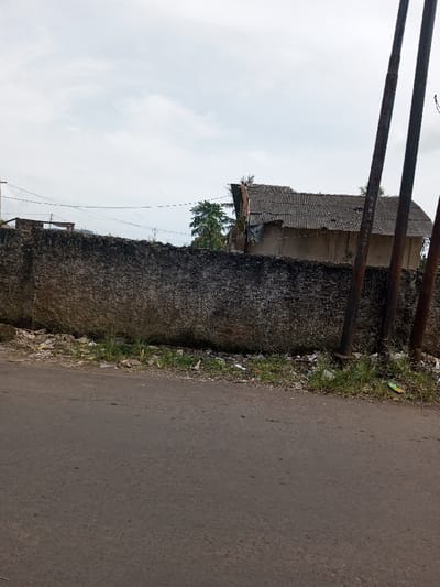 The image depicts a roadside scene in Kandenuwara, Sri Lanka, under an overcast sky. In the foreground, a dark asphalt road spans the lower half of the frame. Immediately adjacent to the road, a narrow, unkempt strip of ground is visible, containing sparse green grass and significant amounts of litter, including plastic bags and various debris. Dominating the midground is a roughly textured, dark grey concrete wall, approximately 1.5 to 2 meters high, running horizontally across the image. The wall exhibits discoloration and signs of weathering. On the right side, two dark, rusted utility poles rise vertically from the ground near the wall. Behind the wall, a dilapidated single-story building is visible on the right, featuring a damaged, corrugated grey roof and light-colored walls. Further left and behind the wall, additional indistinct structures or foundations and green foliage are present. Overhead, multiple thin utility lines stretch horizontally across the frame. No individuals, direct actions, or interactions are visible.