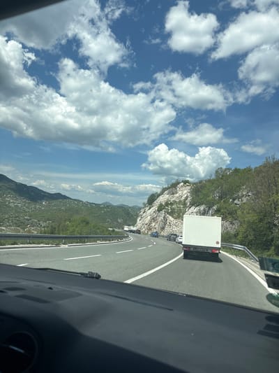 The image presents a daytime view from inside a vehicle traveling on a winding mountain road in Obzovica, Montenegro. The perspective looks forward and slightly to the right, capturing the road curving through scenic, hilly terrain.

Ahead of the viewer's vehicle, a prominent white box truck occupies the right lane of the asphalt road. On its rear, a red circular sticker with the number "80" is clearly visible, denoting a speed limit. The truck's license plate, "06 AT666", is also legible. Behind this truck, several other vehicles, including cars, are queued up, indicating traffic on the road. The viewer's vehicle interior, specifically a portion of the dashboard and windshield frame, is visible in the bottom right corner of the frame.

The road itself is paved with asphalt, featuring distinct white lane markings and a protective metal guardrail along its outer edge. To the left and right of the road, the landscape is characterized by steep, rugged mountainsides covered extensively with lush green trees and shrubs, interspersed with rocky outcrops. In the distance, layers of green mountains and hills stretch under a broad, vibrant blue sky adorned with scattered white, puffy cumulus clouds.

The scene suggests a bright, sunny day, likely during the morning or early afternoon, highlighting a journey through a picturesque, mountainous region. The "06" prefix on the truck's license plate corresponds to the Podgorica region of Montenegro, aligning with the provided location context.