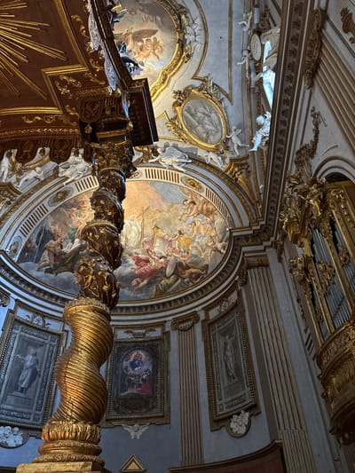 The image depicts the opulent interior of the Cappella del Sacramento (Chapel of the Sacrament) located within the Duomo di San Feliciano (Cathedral of San Feliciano) in Foligno, Italy. The perspective is upward-angled, showcasing the intricate decorations and architecture.
In the left foreground, a prominent, spiraling golden column, part of the high altar's balustrade, rises upwards. Its base features twisted fluting, transitioning to relief carvings resembling foliage or vines higher up. Above this column, a dark, elaborately carved wooden structure with gold accents, possibly a canopy or section of a pulpit, is partially visible.
The central feature is a large, semi-circular ceiling fresco, likely "The Glory of Saints," depicting numerous figures. On the left, a cross is visible amidst robed figures. In the center, a divine figure is surrounded by a radiant light, with angels and saints ascending and adoring. The fresco is framed by a heavily gilded architrave and cornice featuring white stucco cherubs and intricate relief patterns. Above this, the vaulted ceiling segments include smaller frescoes or grisaille paintings within gilded oval frames, interspersed with additional white stucco figures and gold ornamentation.
Below the main fresco, the pale blue-grey walls are adorned with framed artworks. A large, rectangular grisaille-style painting of a standing robed figure is visible on the left wall. Further right, beneath the main fresco, a smaller, colored painting depicting the Madonna and Child with other figures is displayed. Another grisaille-style painting of a robed figure is visible on the right wall. On the far right, a section of a large pipe organ, featuring polished silver pipes set within an extensively gilded wooden structure adorned with scrolling elements, is visible. The entire space is characterized by pervasive gold leaf, detailed frescoes, and white stucco sculptural elements, contributing to an atmosphere of ecclesiastical grandeur. No living individuals are present in the scene.