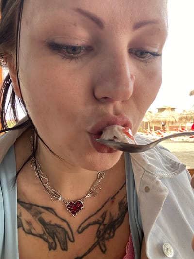A close-up, slightly rotated, ground-level shot depicts a woman eating a dessert with a spoon. The woman has fair skin, green eyes, and dark eyebrows. She is wearing a light blue denim jacket, and visible under the jacket is pink lace fabric. A silver necklace with a red heart-shaped pendant rests on her chest, which also features a dark tattoo of hands. She is eating a white dessert topped with a red sauce. The spoon is silver. The background appears to be outdoors, possibly in Ulcinj, Montenegro, given the awnings and structures in the distance, but this is not definitively identifiable in the provided image.