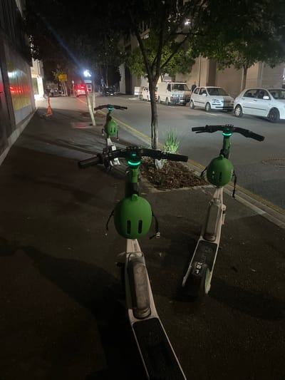 The image shows a nighttime scene on a city street, likely in Brisbane, Australia, with several electric scooters parked on a sidewalk adjacent to a road. The scooters are predominantly green and white, with a green protective helmet on the front of two of them. The scooter closest to the viewer has "Lime" branding visible on its deck. On the road, several cars are parked, including a white van and a white sedan. Streetlights and ambient lighting illuminate the scene. The sidewalk is paved, and there are trees and buildings in the background. A yellow traffic sign is visible on the left side of the image. No people are clearly visible in the foreground, but there appears to be a person in the background near the white van. The overall impression is of a quiet, urban street at night.