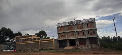 A partly cloudy sky hangs over two buildings. The building on the left is two stories with a wall around it. The building to the right is three stories, with windows and doors appearing unfinished. The buildings stand near a road and are surrounded by vegetation. The phrase "police komba" is visible at the bottom left corner of the image.