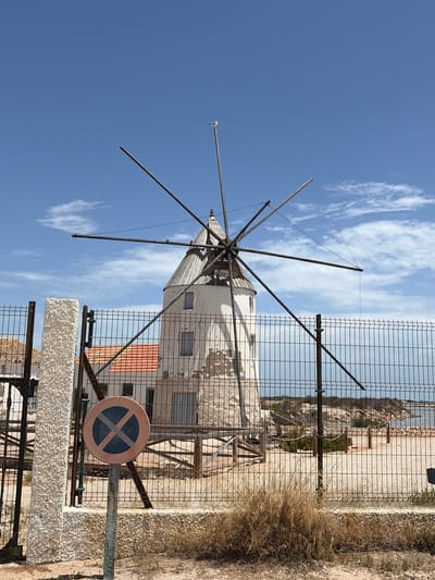 The image depicts a traditional windmill, a historical landmark in the city of San Pedro del Pinatar, Spain, situated near the coast. The windmill, with its whitewashed tower and conical roof, stands behind a metal fence. Its large, wooden sails are extended, though they appear to be made of metal poles in this view. A seagull is perched on one of the sail poles against a bright blue sky with scattered clouds. In the foreground, a circular traffic sign with a blue background and a red border, featuring an "X" symbol, is mounted on a metal post. The surrounding environment includes dry, sparse vegetation, sand, and in the background, a glimpse of the sea and a rocky coastline. The overall scene suggests a sunny day in a coastal area.