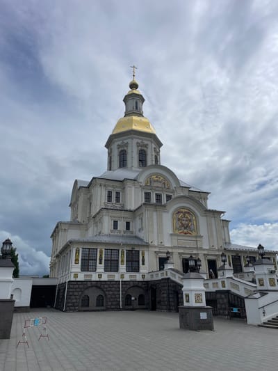 The image shows the Trinity Cathedral in Diveyevo, Russia, a large, white, ornate building with a golden dome topped with a cross. The cathedral is under a cloudy sky. The foreground is a paved courtyard with a red and white barrier. The building has multiple windows and decorative elements, including mosaics on the facade. Some people are visible near the entrance. The architecture suggests a religious or historical site.