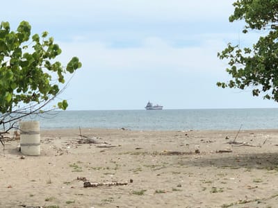 This image depicts a coastal scene in Bangkajaya, Indonesia. In the foreground, a sandy beach is littered with debris, including pieces of wood, plastic, and vegetation. A concrete cylindrical structure is visible on the left side of the beach. In the middle ground, the ocean stretches out towards the horizon, with a large cargo ship visible in the distance. The sky is overcast with scattered clouds. Trees with green foliage frame the left and right edges of the image.