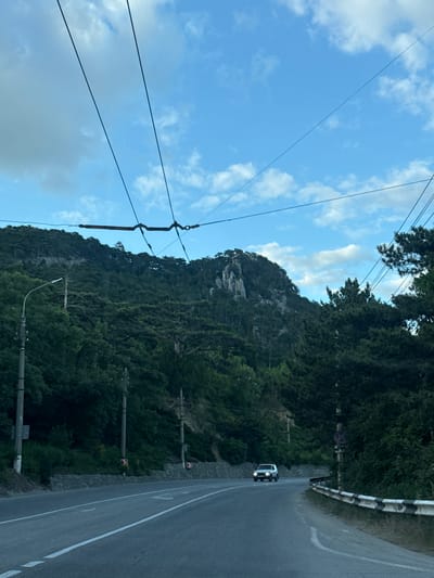 The image depicts a scenic view of a road winding along a steep, forested mountainside under a bright blue sky with scattered clouds. Overhead, power lines crisscross the frame, adding a sense of infrastructure to the natural landscape.

The road itself is paved and has a white dividing line, indicating it's a two-way street. A white vehicle, possibly a van or SUV, is visible on the right side of the road, heading away from the viewer. The mountainside is densely covered in lush green trees, with rocky outcrops visible near the peak.

Along the roadside, there are several utility poles and some signage. One sign appears to be a traffic sign with a red circle and a diagonal line, indicating a prohibition. Another sign has a red and white checkered pattern, likely a warning sign.

The lighting suggests it is daytime, possibly late afternoon given the slight shadow cast by the vehicle. The weather appears clear and pleasant, with the sun shining through the clouds. The overall impression is one of a beautiful, natural environment with a man-made road cutting through it.

There is no visible text in the image, other than what might be present on the signs, which are too small to decipher.