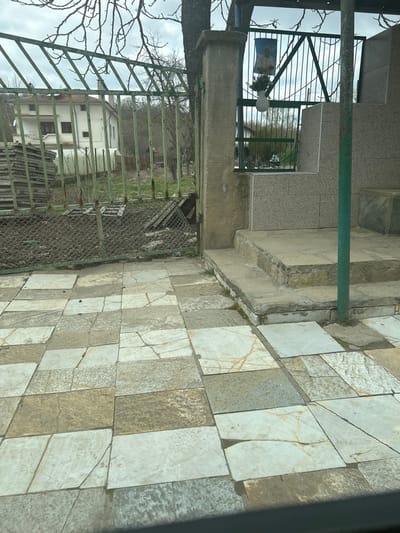The scene depicts an outdoor paved area in Chernodab, Bulgaria, extending towards a fence, a small tiled structure, and a natural background. The foreground consists of an uneven pattern of rectangular and square flagstones in shades of white, beige, and grey, many exhibiting cracks and some dislodged. Small dark debris is scattered on the paving.

To the left, a deteriorated green metal fence with vertical bars and a lower section of damaged wire mesh encloses an unkempt dirt area containing wood scraps and other debris. Behind the fence, a stack of rectangular panels is visible, and further back, a two-story white house with a brown roof and brown window frames stands amidst bare trees.

A thick, vertical concrete pillar, exhibiting etched lines, marks a transition point to the right. Attached to a green metal gate section immediately to the right of this pillar is a poster featuring an elderly man with grey hair and a mustache, wearing a light shirt. Below the poster, a white plastic-like container holding dark, possibly floral, contents hangs from the gate. Adjacent to the concrete pillar on the right is a small, tiled structure or platform with a single step, covered in light grey square tiles. A round green metal pole is positioned in the lower right, partially obscured. The distant background reveals more bare trees under an overcast grey sky. No people are present.