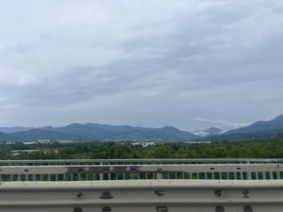 The image captures a panoramic view of the city of Akhalsopeli, Georgia, under a cloudy sky. In the foreground, a metal guardrail of a bridge is visible, with its metallic structure and reflective surface. Beyond the guardrail, a lush green landscape unfolds, characterized by dense trees and vegetation. In the mid-ground, buildings of the city are scattered amidst the greenery, with some appearing to be under construction or development. The background is dominated by a series of majestic mountains, their peaks partially obscured by clouds, creating a dramatic and atmospheric backdrop. The overall scene suggests a tranquil and scenic environment, likely observed from a moving vehicle due to the presence of the guardrail.