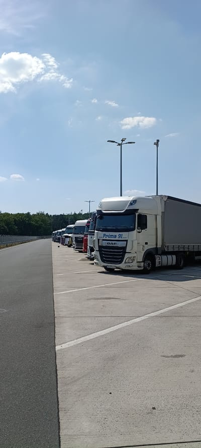This is a wide-angle, vertically oriented photograph of a line of parked semi-trailer trucks on a sunny day in Wachenroth, Germany. The trucks are parked on a concrete lot next to an asphalt road. The sky is bright blue with scattered white clouds. There are two tall streetlights in the foreground, one with a single light and the other with multiple lights. The trucks are white and appear to be in a commercial setting, possibly a rest stop or depot. The closest truck has "Prime 91.com" and "DAF" written on its side. The license plate on this truck is visible and reads "B2976TW". The scene suggests a midday time with clear weather. The overall mood is neutral, depicting a common scene of commercial vehicle parking.