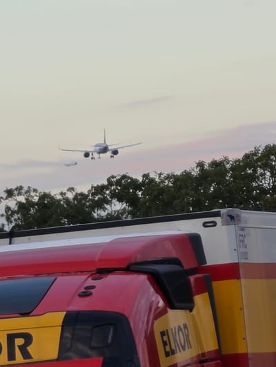 The image shows a red and yellow truck cab in the foreground, with the partial word "ELKOR" visible on a yellow panel. Behind the truck, a line of green trees is visible, and beyond the trees, two airplanes are in the sky. The larger airplane is in the upper center of the image, appearing to be on approach for landing, with its landing gear extended. A smaller airplane is visible to the left and slightly below the larger one. The sky is a pale, hazy color, with hints of pink and orange suggesting either sunrise or sunset. The overall scene appears to be an outdoor location, possibly near an airport or in an area with aerial traffic. The context of Flörsheim, Germany, is not visually evident from the image itself.