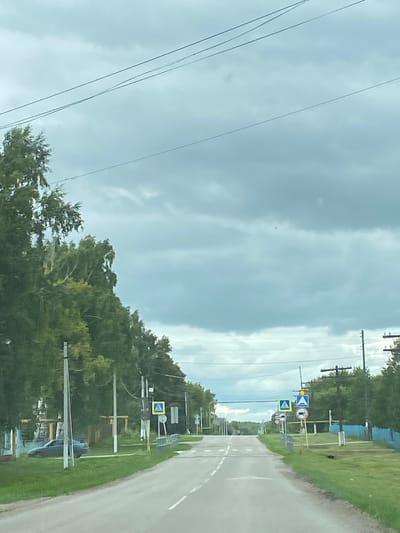 This image depicts a street scene in Nadezhdino, Russia, under a cloudy sky. The road is paved and has white lane markings. On the right side of the road, there are several traffic signs, including a pedestrian crossing sign, a sign indicating a dangerous turn, and a sign prohibiting motorcycles. There are also utility poles with wires running across the scene. Lush green trees line the sides of the road, with some taller trees on the left and a denser line of trees on the right. A blue fence is visible along the right edge of the property. In the foreground, on the left side of the road, a dark-colored car is parked on the grass near some houses. The overall impression is of a quiet rural or semi-rural road.