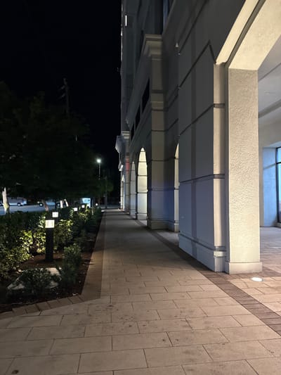 Captured at night, an exterior perspective showcases a building's architectural details. A colonnade structure with evenly spaced pillars runs along the left, leading into the distance. To the right, a paved walkway borders a garden with trees illuminated by small lights. The contrast between the dark night and the lit architecture creates a sense of depth. The image is oriented sideways.