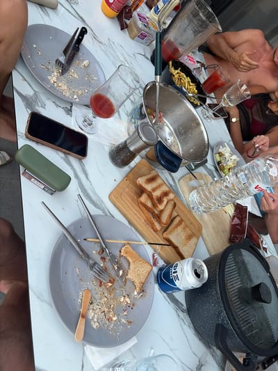 The image depicts a table set for a meal, likely outdoors, with remnants of food and drinks scattered across a marble-patterned surface. Several plates, some with food scraps and utensils, are visible. A glass with a red liquid, likely juice or wine, sits next to a nearly empty glass. A pitcher with a similar red liquid is also present, along with a bowl of French fries and a small salad. Several slices of toasted bread are arranged on a wooden cutting board, along with a can of what appears to be soda or beer. A large bottle of water is also on the table, as is a smartphone and a green case. In the background, a person is partially visible, appearing to be a man with his torso exposed. Another person, a woman, is also partially visible, wearing a dark top. The overall impression is a scene of a recently finished meal, possibly a brunch or lunch, given the presence of toast and fries. The setting appears to be a domestic or hospitality environment, perhaps a patio or balcony. The context of the Municipality of Lefkada, Greece, suggests a warm climate and a leisurely atmosphere.