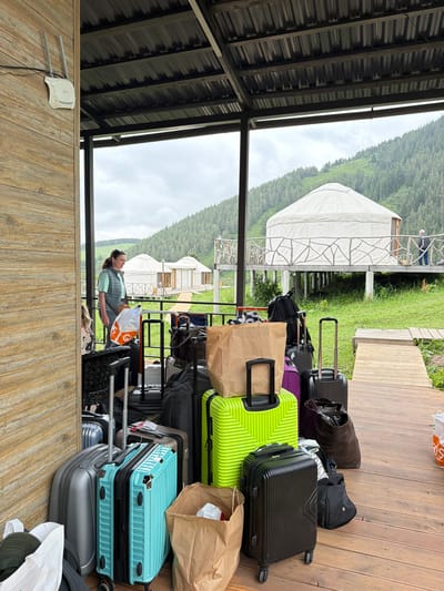 This image depicts a collection of luggage, including several suitcases of various sizes and colors (turquoise, lime green, black, silver), as well as bags and backpacks, arranged on a wooden deck. A woman wearing a light blue vest over a mint green shirt is standing next to some of the luggage, looking towards the right side of the frame. In the background, there are several white yurt-like structures situated on stilts, surrounded by lush green grass and a backdrop of a tree-covered mountain range under a cloudy sky. A Wi-Fi router is mounted on a wooden wall to the left of the frame. The setting appears to be a resort or glamping site in the vicinity of Kaindy, Kyrgyzstan, potentially near the Kaindy Gorge, known for its unique submerged forest.