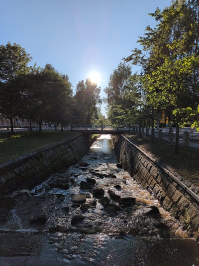 The image captures a sunny day with the sun visible and shining brightly in a clear blue sky, positioned towards the top center. The scene depicts a canal or narrow river with stone-lined banks, flowing through what appears to be a park or urban green space in Oulu, Finland. The water is shallow, with numerous rocks visible within it, and the flow appears somewhat turbulent. Trees line the banks on both sides, with dense foliage casting shadows. A bridge crosses the canal in the mid-ground, with a paved walkway or road visible beyond it, bordered by trees and streetlights. The overall impression is of a well-maintained urban environment on a pleasant day. There are no people visible in the image.
