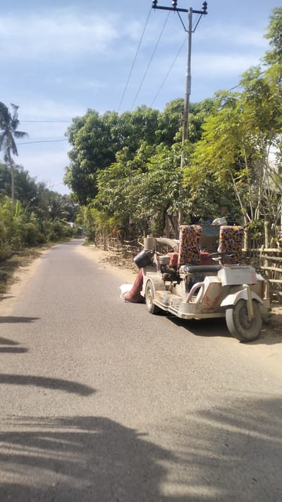 A three-wheeled vehicle, possibly a motorized tricycle, is parked on the side of a paved road. The vehicle is white and has a canvas-covered seat with a floral pattern. It appears to be modified for some kind of work, with visible engine components and a large pipe extending from the side. The road is bordered by vegetation, including trees and bamboo fencing. In the background, a utility pole with power lines is visible, and a cluster of houses can be seen further down the road. The sky is blue with scattered clouds. The lighting suggests it is daytime. The scene is located in Meunasahkrueng, Indonesia.