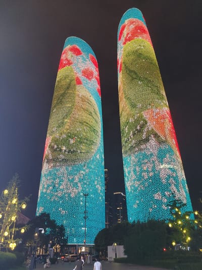 The image depicts two illuminated skyscrapers at night in Wuhou District, China. The buildings are covered in a pattern of lights that form abstract designs in various colors, predominantly teal, yellow, and red. The lighting on the towers appears to depict a stylized natural scene, possibly fruit like watermelons and floral elements. In the foreground, a Christmas tree adorned with yellow lights stands to the left of the frame. Further to the right and behind the tree, there is foliage and streetlights. Several people are visible on a walkway in front of the buildings, some walking and others appearing to be standing or sitting. Cars are also present on the street. The sky is dark, indicating nighttime.