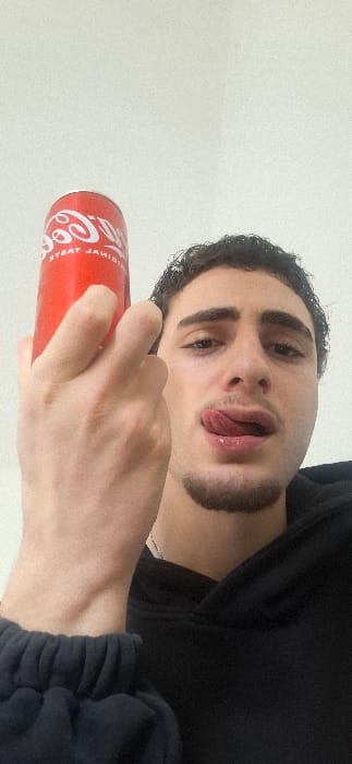 A young adult male is depicted in a close-up, vertically oriented photograph, positioned against a plain, light-colored background. He has dark, curly hair, dark eyes, and light stubble on his chin and upper lip. He is wearing a dark-colored, possibly black, hooded garment, with the ribbed cuff of an additional dark sleeve visible in the lower left. A thin, silver-colored chain is discernible around his neck. His left hand, with visible knuckles, holds a red Coca-Cola can vertically in front of the upper left portion of his face. The can's label features the inverted "Coca-Cola" logo and "ORIGINAL TASTE." The male's facial expression shows his tongue extended to touch the corner of his mouth, with his lips slightly parted and appearing moist. The photograph was taken in Saint-Martin-d'Hères, France.